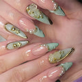 A close-up of a hand showcasing elaborate, long stiletto-style press-on nails. The nails feature a glossy pale pink base with a mint green leaf design on the tips, decorated with 3D white roses, gold foil details, and small white faux pearls, Obroi