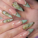 A close-up of a hand showcasing elaborate, long stiletto-style press-on nails. The nails feature a glossy pale pink base with a mint green leaf design on the tips, decorated with 3D white roses, gold foil details, and small white faux pearls, Obroi