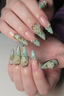 A close-up of a hand showcasing elaborate, long stiletto-style press-on nails. The nails feature a glossy pale pink base with a mint green leaf design on the tips, decorated with 3D white roses, gold foil details, and small white faux pearls, Obroi