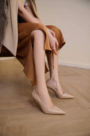A person seated indoors wearing nude beige pointed-toe slingback pumps with a matching beige draped wrap, against a light neutral wall and wood floor, Obroi