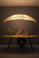 Person sitting at a table with a large feather-shaped lamp above, on a dark background, Obroi