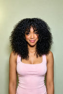 A woman with voluminous, curly black hair wearing a light pink ribbed tank top, standing against a solid pale sage green background, Obroi