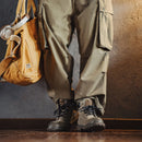 Person wearing brown pants and boots with a brown bag on a wooden floor, Obroi