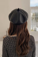 A person viewed from behind wearing a black faux-leather beret hat, with long brown hair and a black-and-white patterned sweater, standing indoors near a window, Obroi