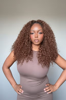 A woman with a full, voluminous curly wig in a warm brown shade, wearing a mauve-brown top, posing indoors, Obroi