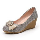 A women's wedge pump shoe featuring a gray and white textured tweed upper, a beige glittery bow, and a jeweled rhinestone brooch on the toe. The shoe has a tan wedge heel and a matching tan outsole, shown against a plain white background, Obroi