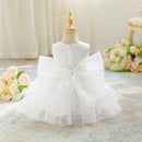 A white toddler girl's party dress displayed on a wooden dress form mannequin. The dress features a sleeveless bodice, a large layered ruffle bow on the back, and a tiered tulle skirt, Obroi