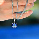 Silver necklace with an evil eye pendant held by a hand against a blue background, Obroi