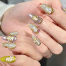 Close-up of manicured fingering with long almond-shaped nails featuring a mix of lavender glitter, white floral patterns, gold foil details, and one bright chartreuse nail, Obroi