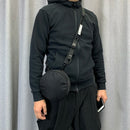 Person wearing a black outfit with a round black bag against a gray curtain background, Obroi