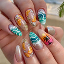 A close-up of a hand with decorated press-on nails, set against a blurred beach background. The nails feature tropical designs including orange slices, teal waves, a pink sunset with a palm tree, and metallic glitter, Obroi