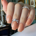 Hand with peach-colored nails and silver rings on a blurred indoor background, Obroi