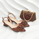 A pair of women's pointed-toe slingback flats in a glittery bronze-brown finish with layered fabric rose embellishments on the toes, shown alongside a matching glittery clutch handbag with a gold-tone chain clasp on a white wavy studio background, Obroi
