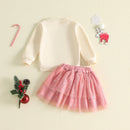 A children's two-piece outfit set featuring a cream long-sleeve sweatshirt and a pink tulle skirt with silver glitter, styled flat-lay with holiday decorations on a light beige background, Obroi