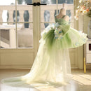 A light green and white tulle formal dress displayed on a mannequin in a bright room with large windows and floral decor, Obroi