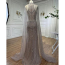 A floor-length beaded evening gown displayed on a mannequin in an indoor room with paneled walls, wood flooring, and sheer curtains. The dress has a fitted silhouette with extensive beadwork and a long sheer train, Obroi