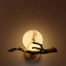 A wall-mounted decorative light fixture featuring a glowing oval moon-like bulb with a tree branch silhouette in front, mounted on a round base against a warm brown wall, Obroi