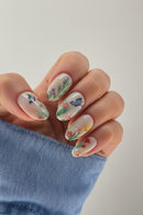 Nails with floral and butterfly design held by a hand wearing a blue sleeve, Obroi