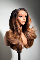 A long, wavy wig with dark brown roots and warm caramel-brown highlights, styled on a mannequin bust against a plain light gray background, Obroi