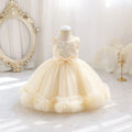 A cream-colored toddler girl's party dress displayed on a mannequin in a bright, elegant room. The dress features a sleeveless lace bodice, a matching satin bow at the waist, and a voluminous tulle skirt with ruffled trim, Obroi