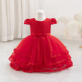 A bright red toddler girl's party dress displayed on a white mannequin dress form. The dress features a textured honeycomb pattern bodice, short puff sleeves, a matching red satin bow at the waist, and a tiered tulle skirt with embroidered lace trim, Obroi