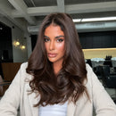 A woman with long, wavy, dark brown hair is shown from the chest up, wearing a light beige cardigan over a white top. The background is a modern salon interior with salon chairs and bright ceiling lights, Obroi