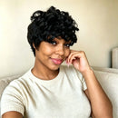 A woman with a short, curly black wig wearing a cream-colored short-sleeve knit top, posing on a light beige couch against a neutral wall, Obroi