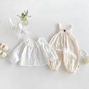 A baby girl's two-piece outfit set featuring a white long-sleeve blouse with a ruffled collar and a cream crinkle-textured romper with suspenders, styled in a flat lay with baby accessories on a light gray background, Obroi