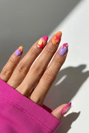 A close-up of a hand with colorful, stylized almond-shaped nail art. The nails feature gradients and abstract designs in pink, orange, purple, yellow, and white, with a bright pink sleeve visible at the bottom left against a neutral gray background.