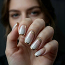 A close-up of a hand with manicured nails painted in a white-to-pale-pink gradient with metallic gold glitter and gold French tips, with a blurred face in the background, Obroi