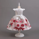 A white girl's party dress displayed on a white mannequin dress form against a solid gray studio background. The dress features a white sleeveless bodice with short flutter sleeves, a matching white satin waistband and bow, and a full white skirt printed with a vibrant red and pink floral pattern, Obroi