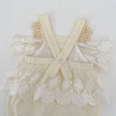 A cream-colored baby romper with white floral lace trim, featuring wide adjustable shoulder straps and a ruffled tulle skirt, displayed against a textured white wall, Obroi