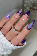 A close-up of a hand with long stiletto-shaped nails featuring a lavender-to-pink gradient and dark purple glossy sections, each decorated with small gold glitter accents. The hand is wearing a white ribbed sleeve and a gold ring with clear stones, Obroi