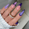 A close-up of a hand with long stiletto-shaped nails featuring a lavender-to-pink gradient and dark purple glossy sections, each decorated with small gold glitter accents. The hand is wearing a white ribbed sleeve and a gold ring with clear stones, Obroi