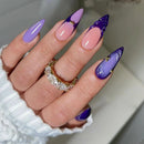A close-up of a hand with long stiletto-shaped nails featuring a lavender-to-pink gradient and dark purple glossy sections, each decorated with small gold glitter accents. The hand is wearing a white ribbed sleeve and a gold ring with clear stones, Obroi