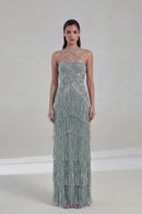 A model wearing a floor-length, strapless gown with crisscross halter-style straps and cascading fringe embellishments in a pale sage green and silver tone, set against a neutral studio backdrop with the brand name Obroi in the top left corner.
