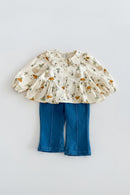 A toddler girl's two-piece outfit featuring a cream long-sleeve blouse with a green and orange woodland animal print and ruffled collar, paired with blue denim cropped pants, laid flat on a light gray background, Obroi