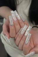A close-up of hands showcasing long, coffin-shaped acrylic nails decorated with intricate white butterfly designs, silver rhinestone borders, and small gold glitter accents, Obroi