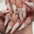 A close-up of manicured fingernails with long almond-shaped tips, decorated with white lace, pink roses, glossy white bows, and small rhinestone accents, shown on a hand, Obroi