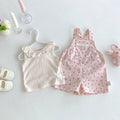 A baby girl's two-piece outfit set featuring a cream ruffled tank top on a pearl hanger and a pink floral overall romper, styled in a flat lay with baby accessories on a white background, Obroi