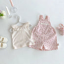 A baby girl's two-piece outfit set featuring a cream ruffled tank top on a pearl hanger and a pink floral overall romper, styled in a flat lay with baby accessories on a white background, Obroi