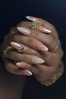 A close-up of a hand showcasing long, almond-shaped artificial nails with various decorative designs including white polka dots, pearl accents, and textured swirls. The nails are styled with multiple gold and silver rings against a dark, solid background, Obroi