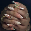 A close-up of a hand showcasing long, almond-shaped artificial nails with various decorative designs including white polka dots, pearl accents, and textured swirls. The nails are styled with multiple gold and silver rings against a dark, solid background, Obroi