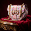 A pink shoulder bag with a multicolor iridescent snakeskin pattern, gold chain straps, and a jeweled rhinestone embellishment on the front flap, displayed on a red velvet draped table, Obroi