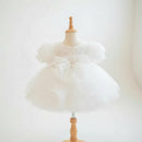 A white toddler girl's party dress with puffy sleeves and a large bow at the waist, displayed on a wooden dress form mannequin, Obroi