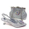 A silver rhinestone-embellished slingback high heel shoe shown in front of a matching silver rhinestone clutch handbag with a floral crystal embellishment and chain strap, on a white background, Obroi