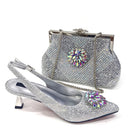 A silver rhinestone-embellished slingback high heel shoe shown in front of a matching silver rhinestone clutch handbag with a floral crystal embellishment and chain strap, on a white background, Obroi