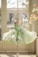 A light green children's party dress displayed on a mannequin in front of a bright window. The sleeveless dress features a fitted embellished bodice and a voluminous tulle skirt decorated with large 3D floral appliqués in pale green, white, and soft blue, Obroi