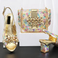 A matching women's fashion set by Obroi, featuring gold metallic slingback high-heeled sandals with jeweled embellishments and a matching iridescent snakeskin-patterned handbag with gold hardware, displayed on a white block against a white background, Obroi