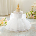White toddler girl's party dress displayed on a wooden dress form mannequin, featuring a sleeveless bodice with floral applique, a tiered tulle skirt, and a large bow on the back, set against a soft cream curtain background with decorative flowers, Obroi